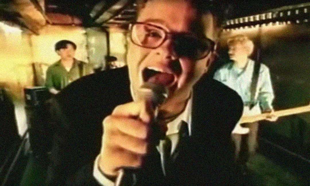 A man in glasses passionately sings into a microphone in the foreground, with two other musicians playing guitar in the background under warm lighting in what appears to be a music video setting.
