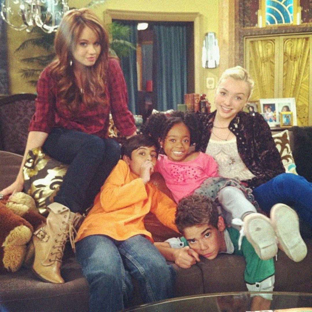 Five children and teenagers sit and lounge playfully together on a couch in a cozy living room, smiling and appearing to enjoy each other's company. The background features warm decor and a decorative mirror.