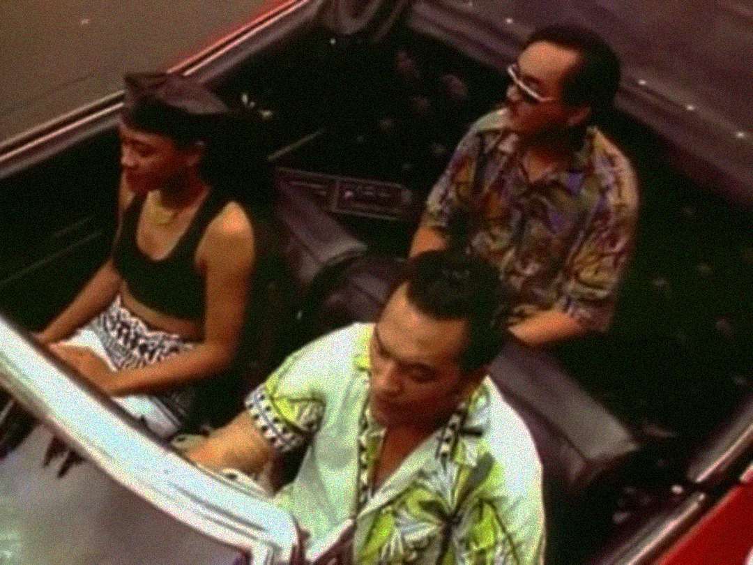 Three people ride in a red convertible car. The driver and passenger in the back are men in patterned shirts; the front passenger is a woman in a black top and headscarf. The image has a vintage, grainy quality.