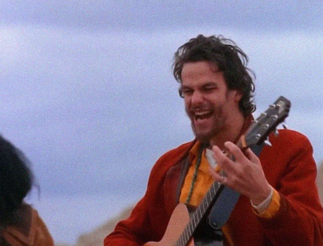 A man with messy hair and a beard, wearing a red jacket, joyfully plays an acoustic guitar outdoors under a cloudy sky.