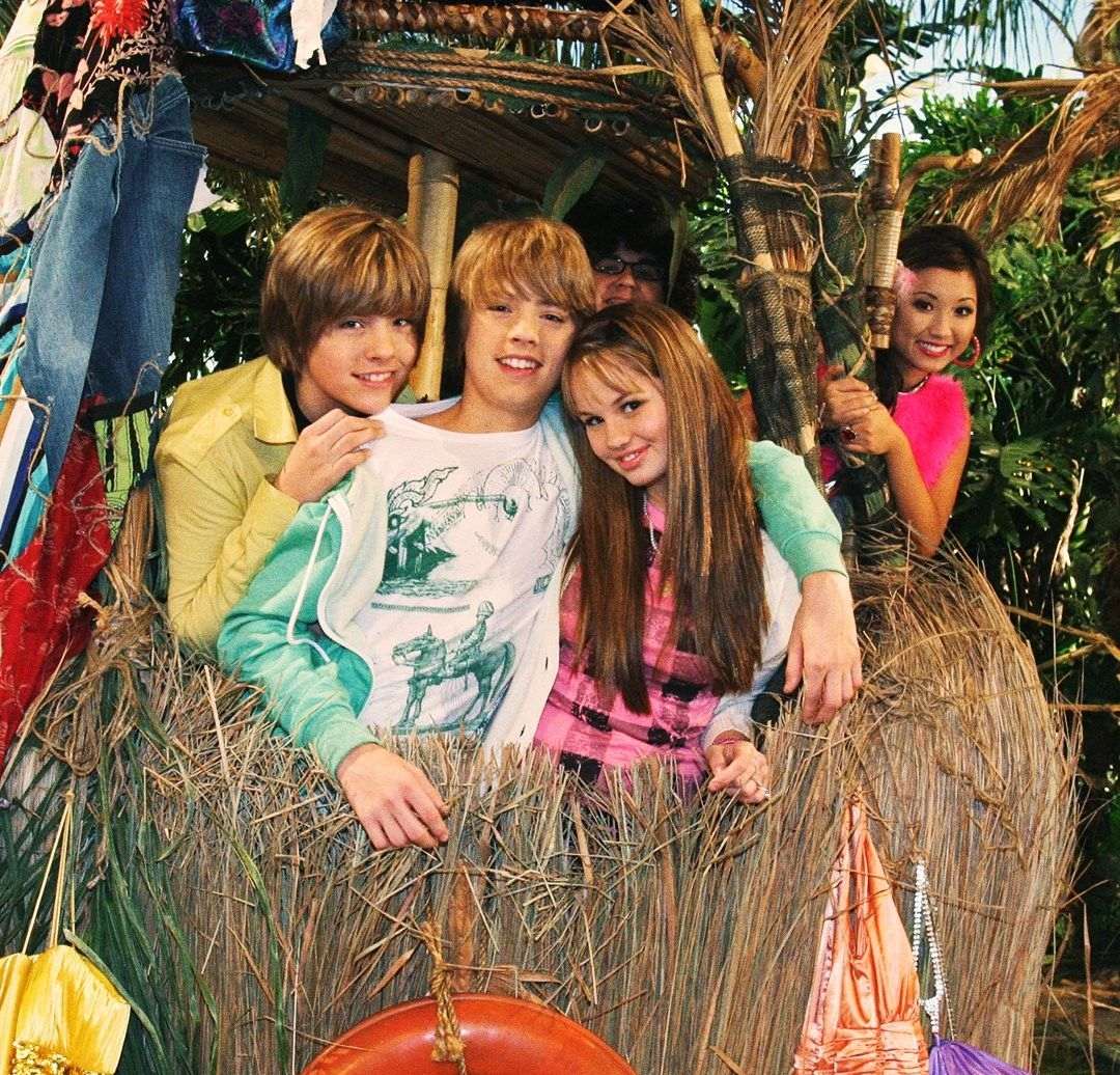 Three teenagers lean on a thatched hut, smiling, with another teen partially visible behind them. A girl in bright pink stands at the edge, waving and smiling. Tropical plants surround the group.