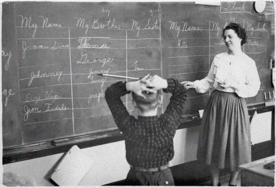 A teacher smiles while standing by a chalkboard with students’ names written under headings like “My Brother,” “My Sister,” and “My Name.” A young boy stands in front, facing the board with his hands on his head.