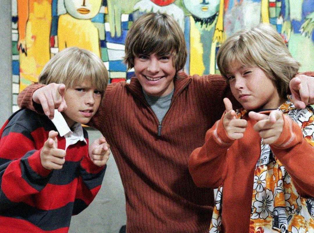 Three smiling boys pose playfully in front of a colorful mural. The two on the sides are wearing striped sweaters and making finger guns, while the one in the middle wears a brown sweater and points toward the camera.