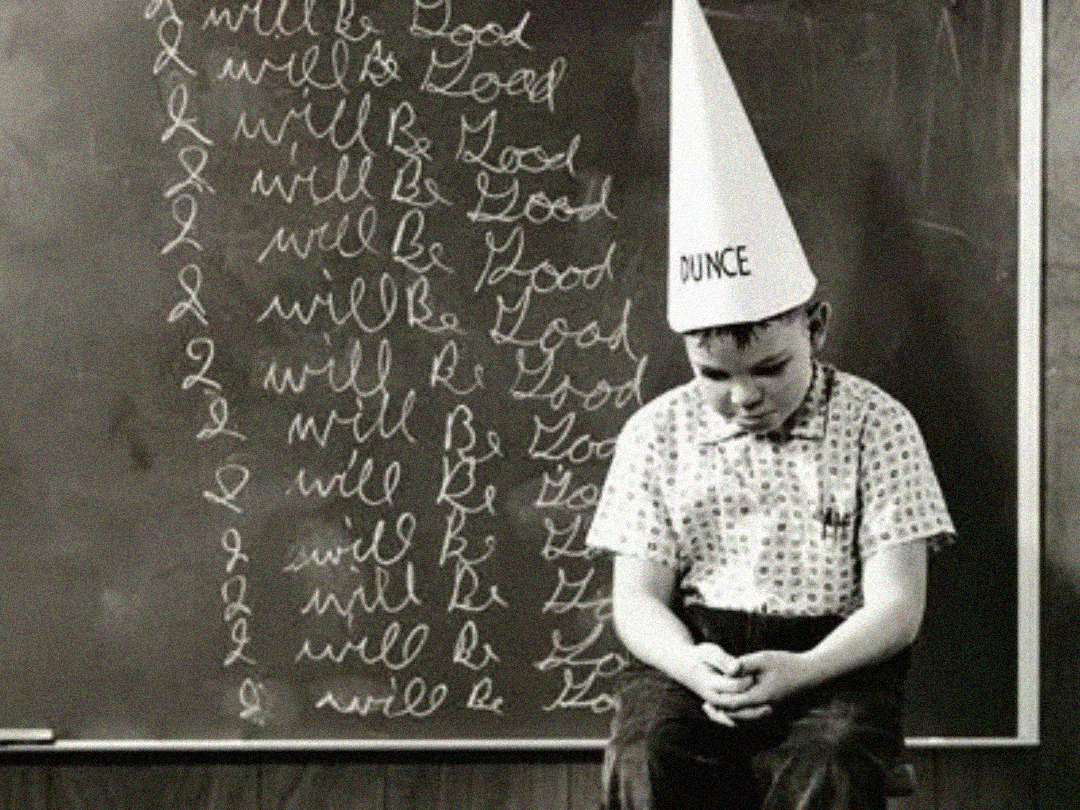 A boy wearing a "dunce" cap sits on a stool in front of a chalkboard. The chalkboard is repeatedly written with the phrase "I will be good." The boy looks down with a sad expression.