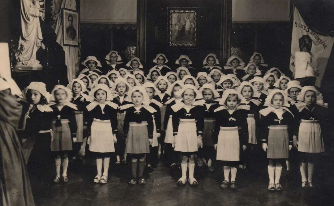 A vintage black-and-white photo of a group of young girls in uniform with white collars and bonnets, standing in rows indoors for what appears to be a formal event or ceremony.