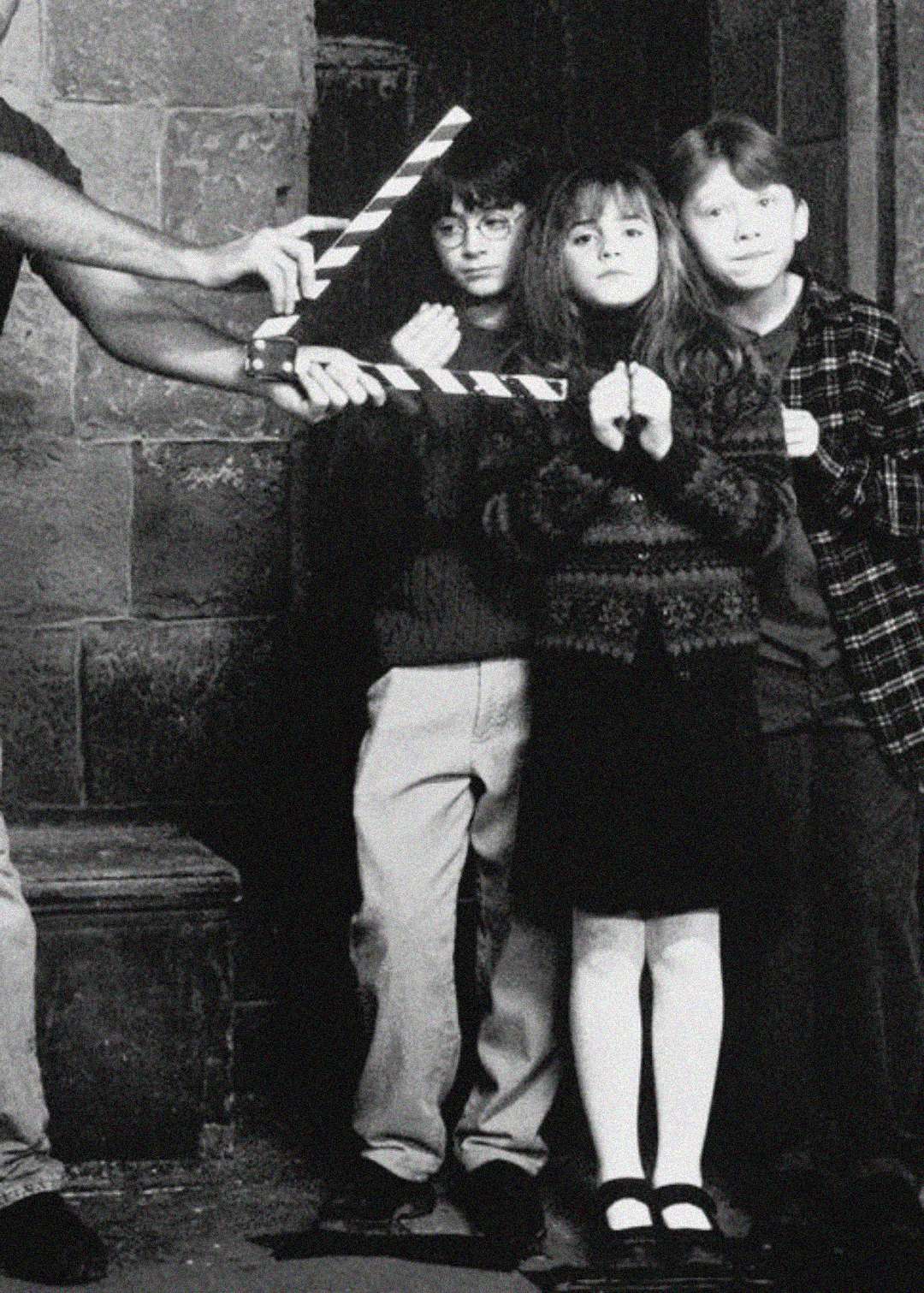 Three children stand closely together on a film set, looking serious. A crew member holds a clapperboard in front of them. The image is in black and white and has an old, behind-the-scenes atmosphere.