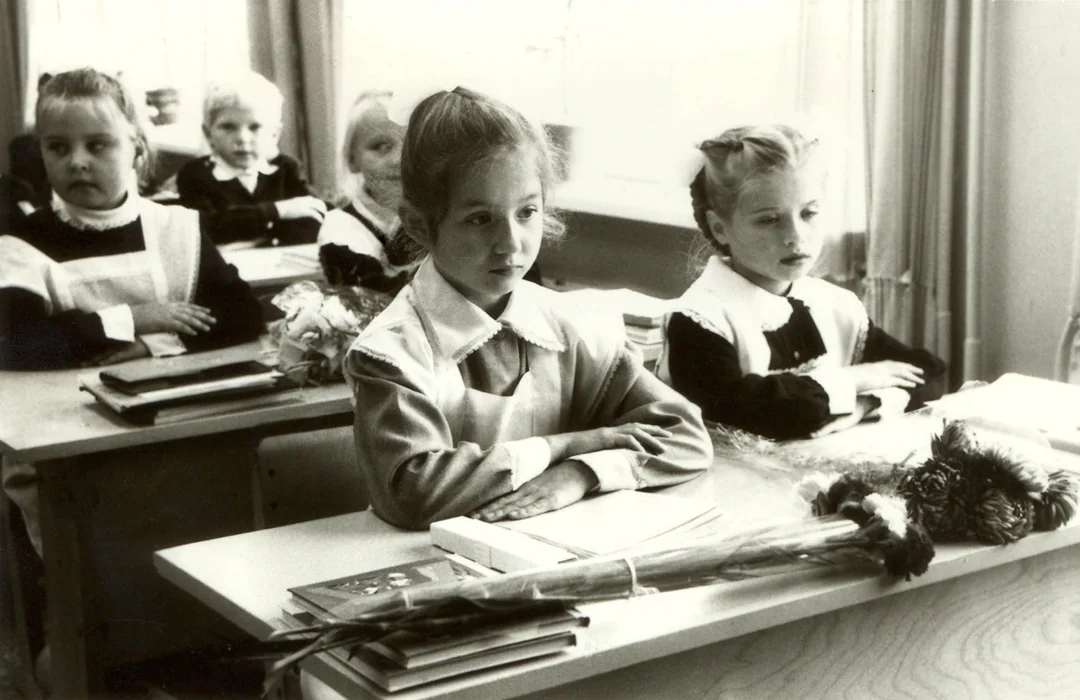 Black and white photo of young girls in school uniforms sitting at desks in a classroom. Books and flowers are on the desks, and sunlight streams through the window behind them.
