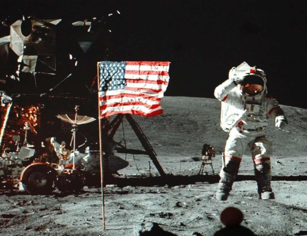 An astronaut in a spacesuit stands on the Moon next to an American flag, with lunar equipment and the lunar module in the background against a dark sky.