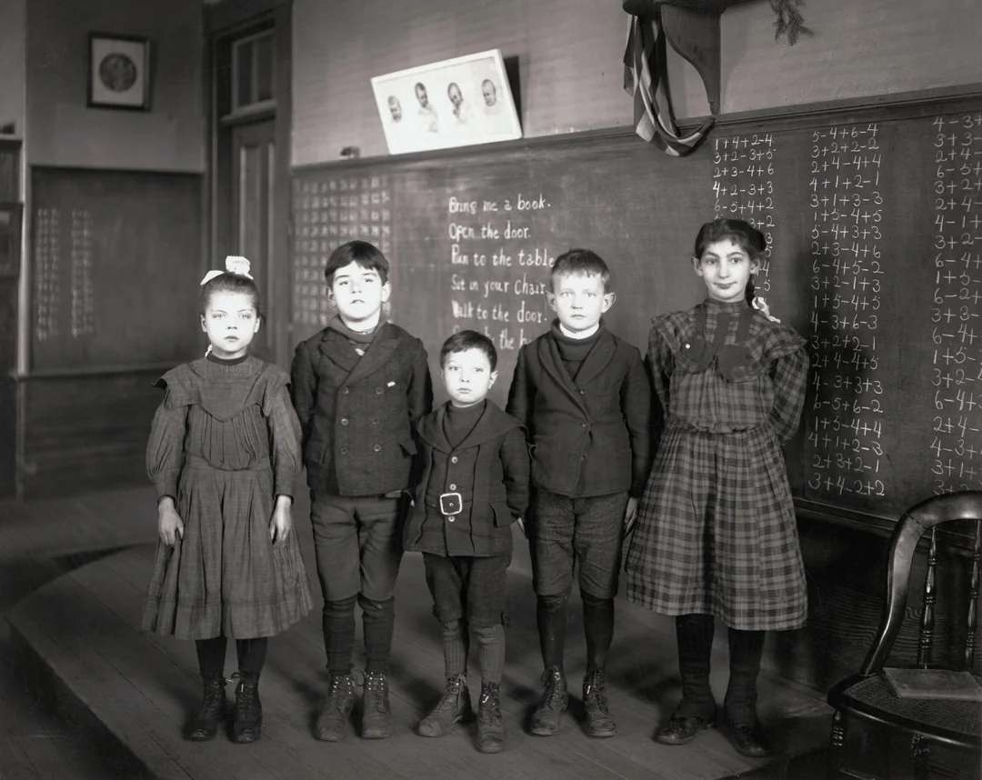 Five children in old-fashioned clothing stand in a classroom, posed in front of chalkboards with arithmetic problems and instructions written on them. The classroom appears to be from the early 20th century.