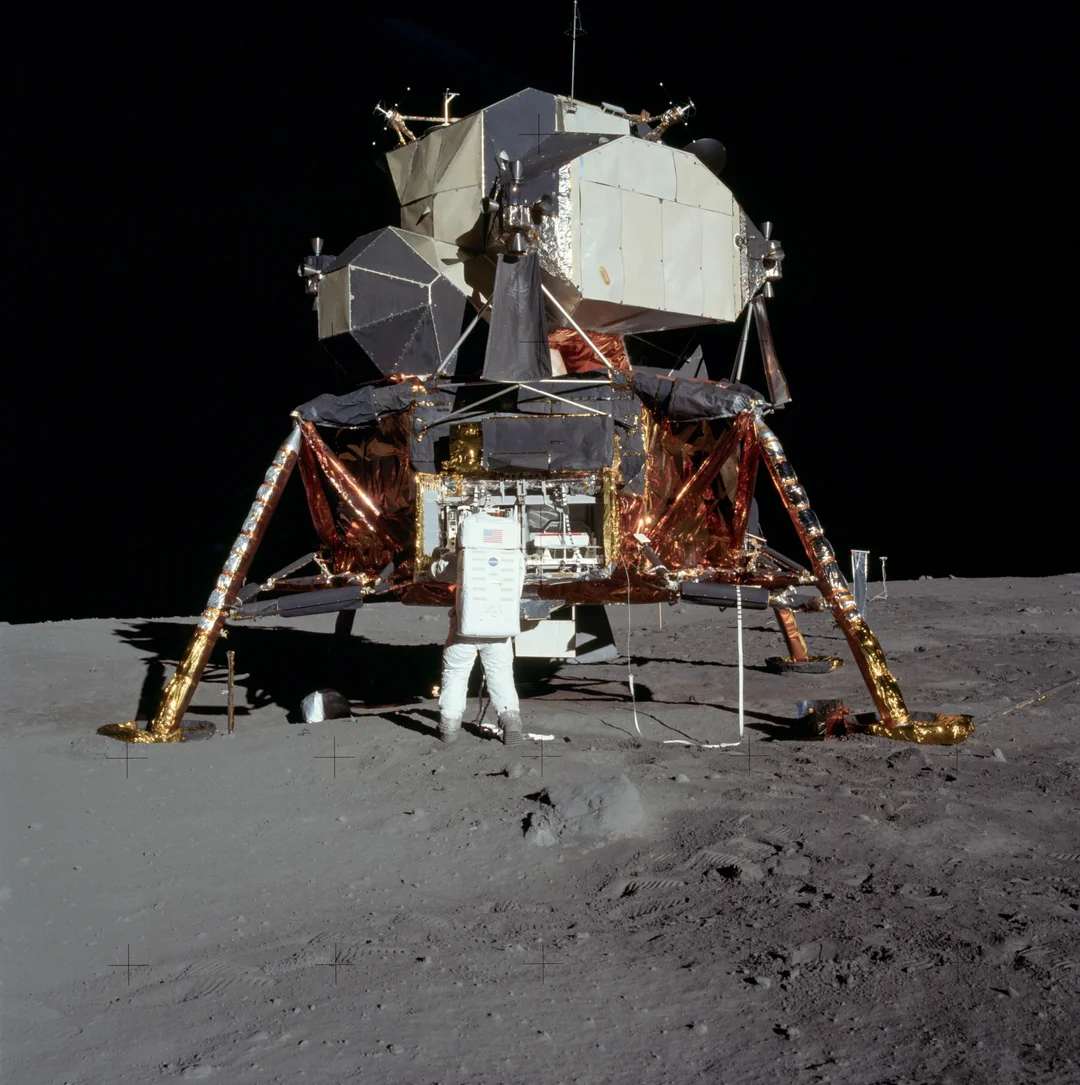 An astronaut stands next to the Apollo Lunar Module on the moon’s surface, with the lunar lander’s metallic structure and legs visible against the dark sky and gray, dusty lunar ground.