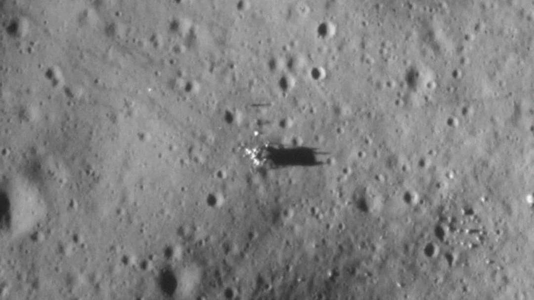 A black-and-white aerial image of a cratered lunar surface shows a small object casting a dark shadow, surrounded by scattered debris and impact marks.