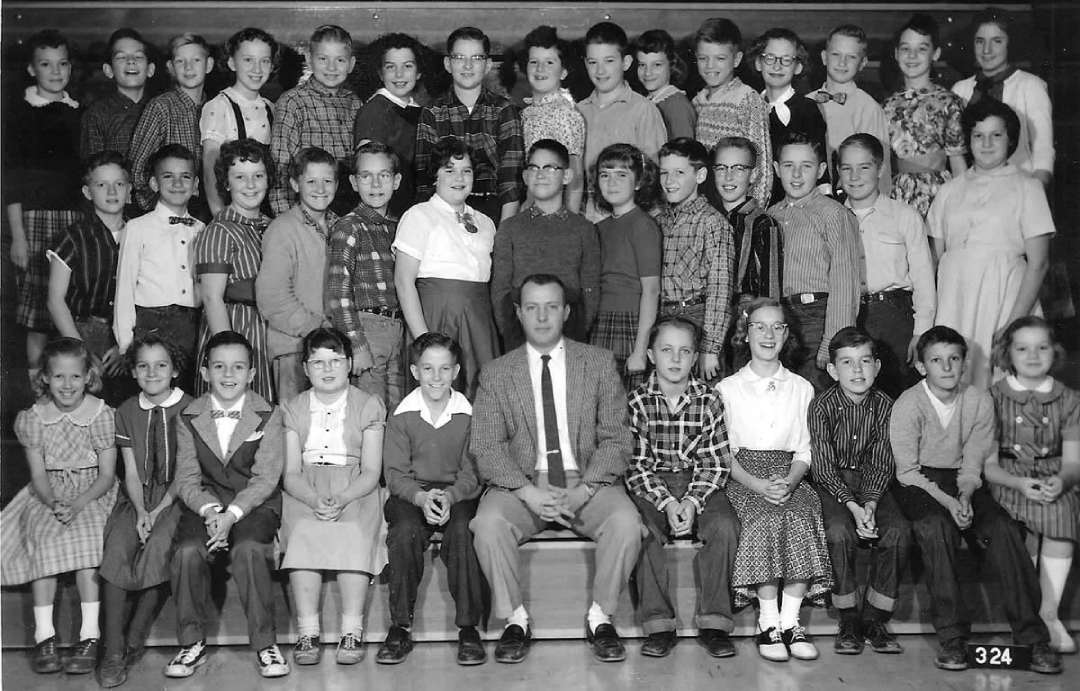 A black and white photo of a school class with young children and one adult male teacher sitting in the center of the front row. The children are posed in four rows, smiling and dressed in 1950s clothing.