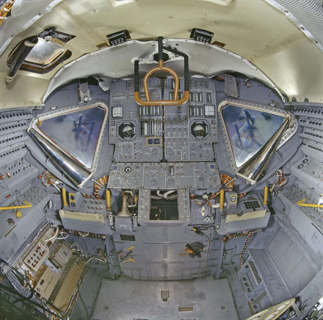 The image shows the control panel and cockpit interior of a spacecraft, with numerous switches, gauges, buttons, and two triangular windows. The space is compact and filled with intricate instrumentation.