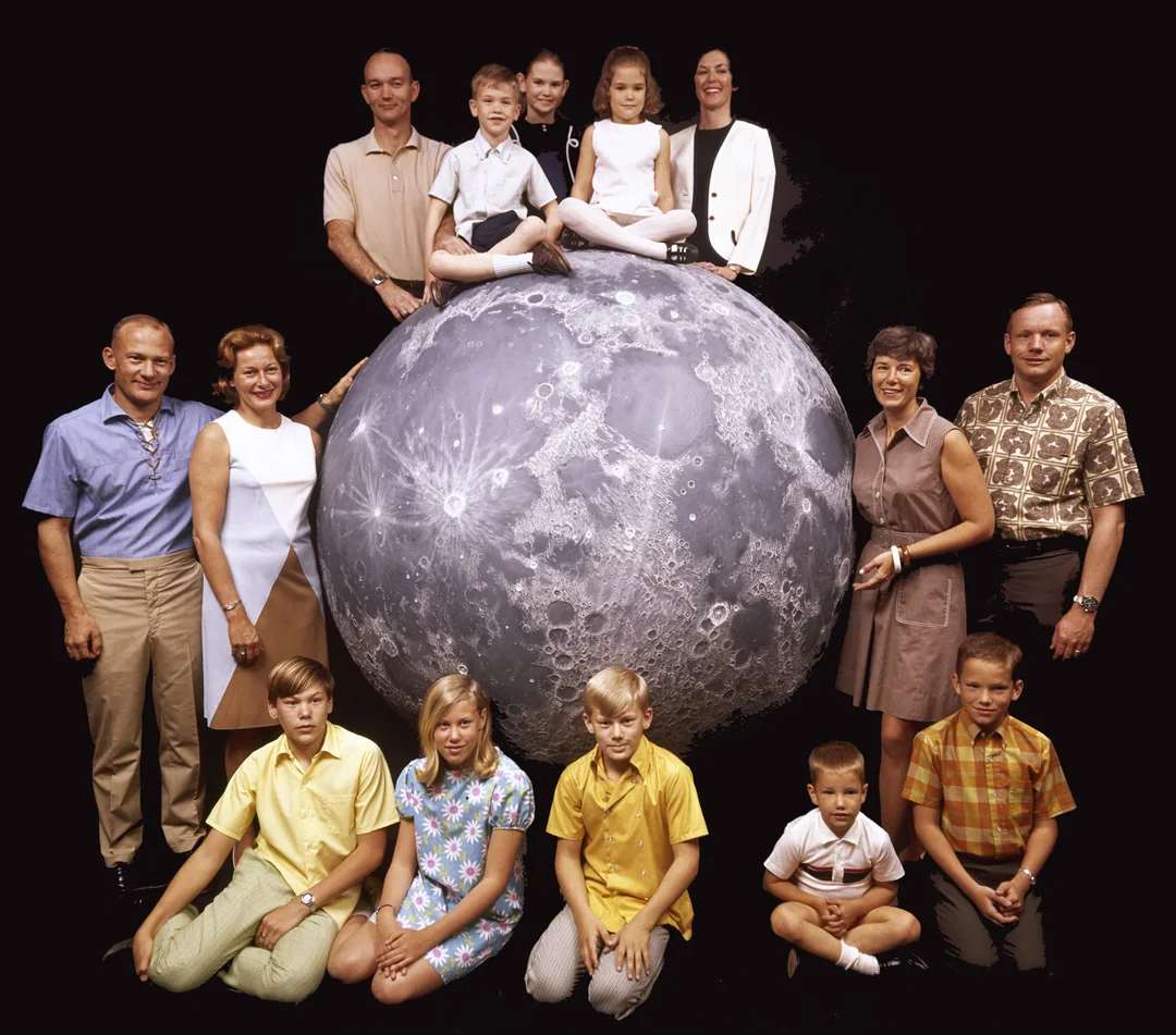 A group of adults and children pose around and on top of a large model of the Moon, with a black background. Some children sit on the Moon; the group smiles for the camera.