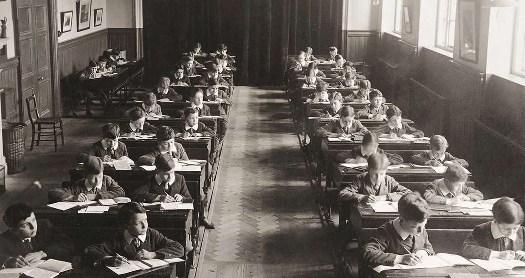 Black and white photo of children sitting at individual desks in neat rows, writing in exercise books in a classroom with wooden floors and large windows. A teacher sits at the back, observing.