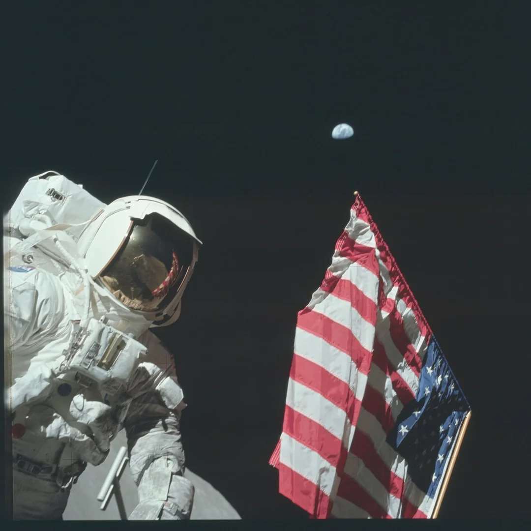 An astronaut in a spacesuit stands on the Moon next to a U.S. flag, with the Earth visible in the dark sky in the background.