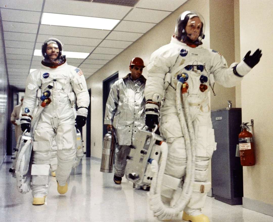 Two astronauts in white space suits walk down a hallway, carrying equipment, followed by a person in a silver suit. The astronauts have helmets on and look focused. A fire extinguisher is mounted on the wall to the right.