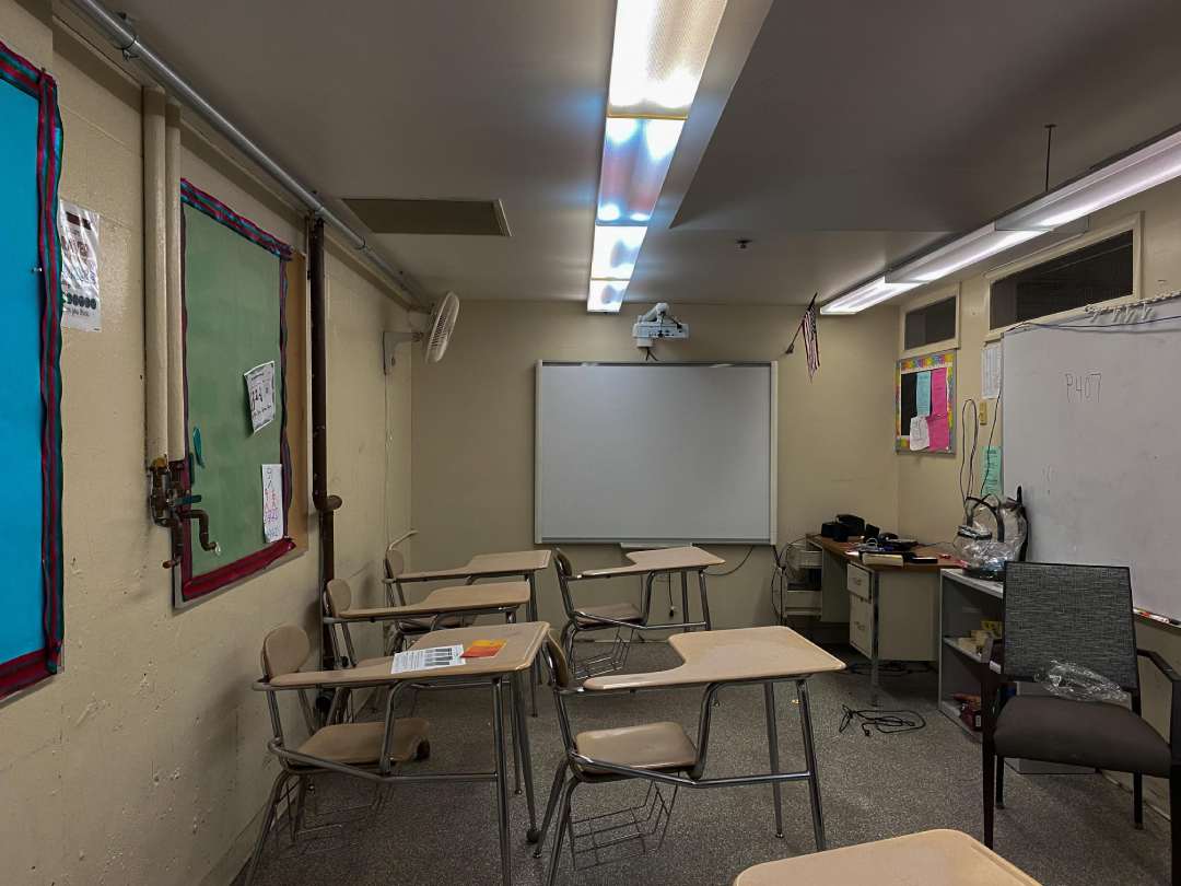 A classroom with empty desks and chairs, bulletin boards on the walls, a whiteboard at the front, and a teacher’s desk with various items. Fluorescent lights are on, and the room appears unoccupied.