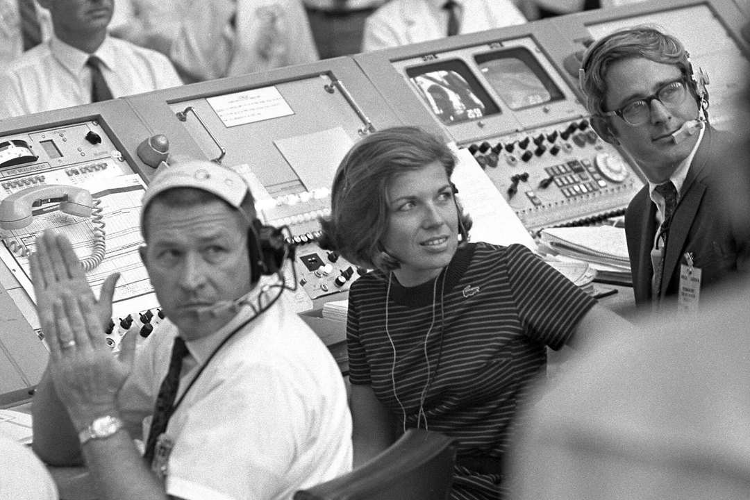 Three people wearing headsets sit at a console filled with buttons and monitors in a busy mission control room, watching intently. The atmosphere is focused and professional.