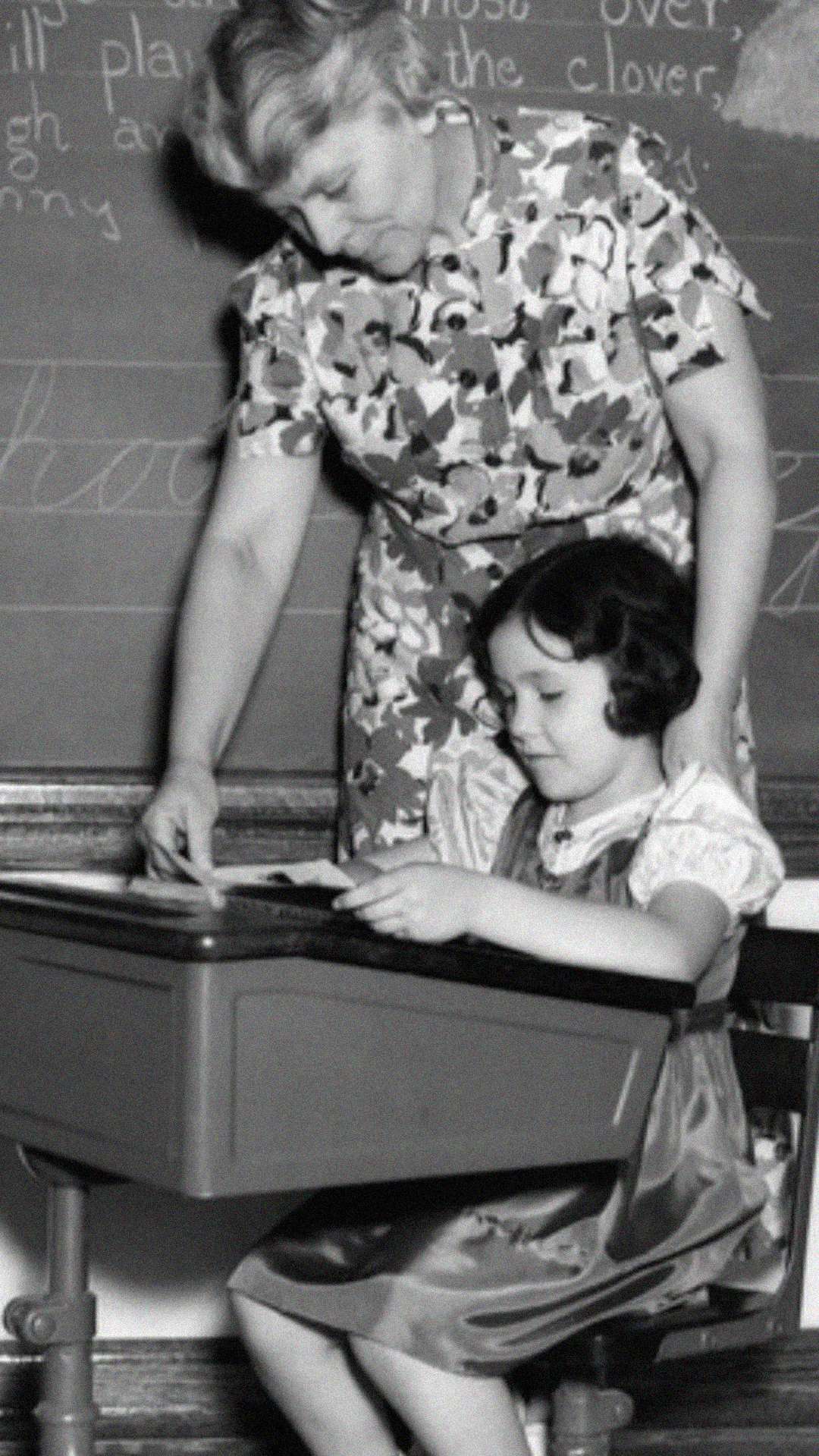 A woman stands beside a young girl seated at a school desk, helping her with her work. Both are focused, and a chalkboard with writing is visible in the background. The scene appears to be from a classroom in the past.