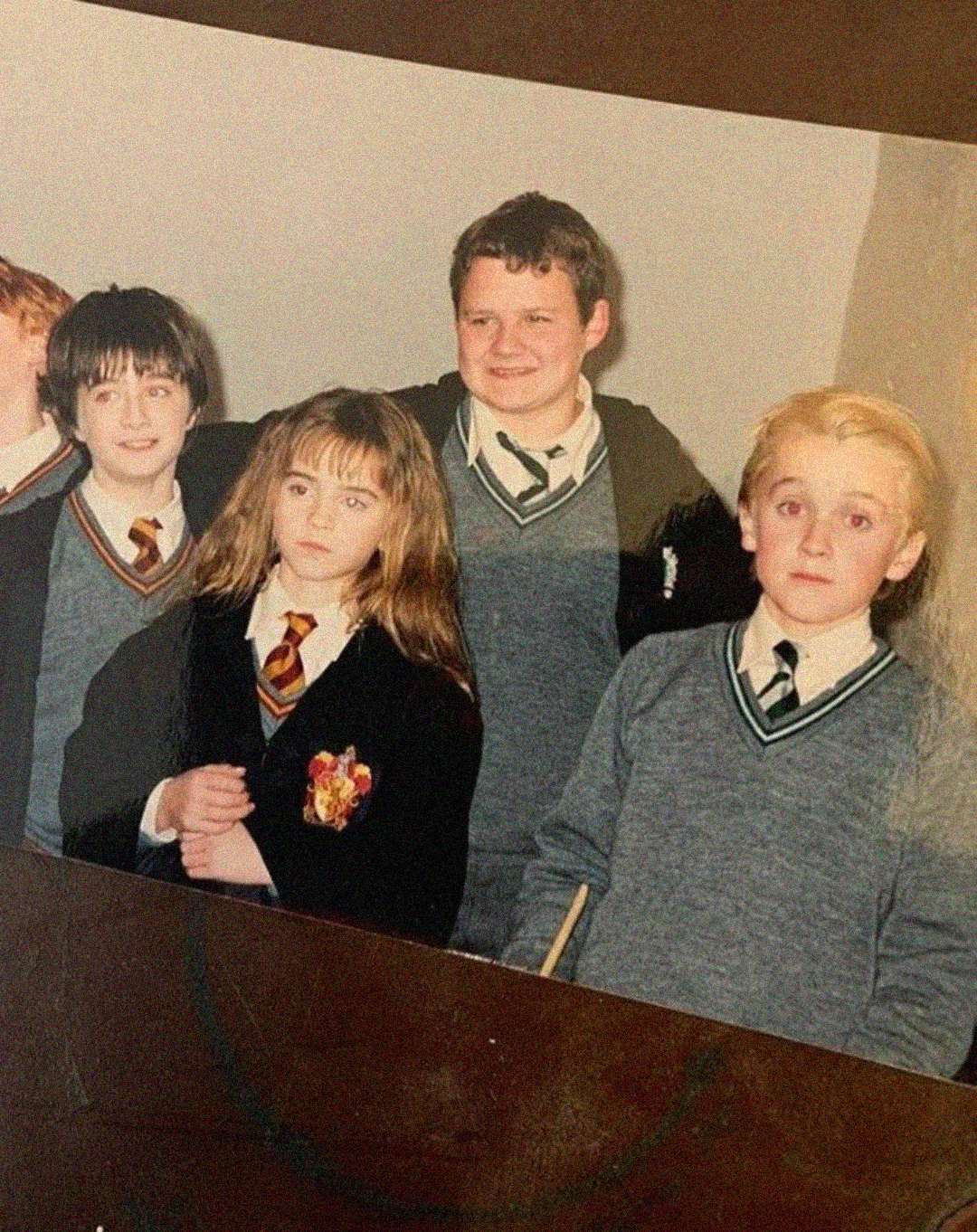 Four children wearing school uniforms, including ties and sweaters, stand together indoors. Three have dark hair, and one has light blond hair. They appear to be posing for a group photo.