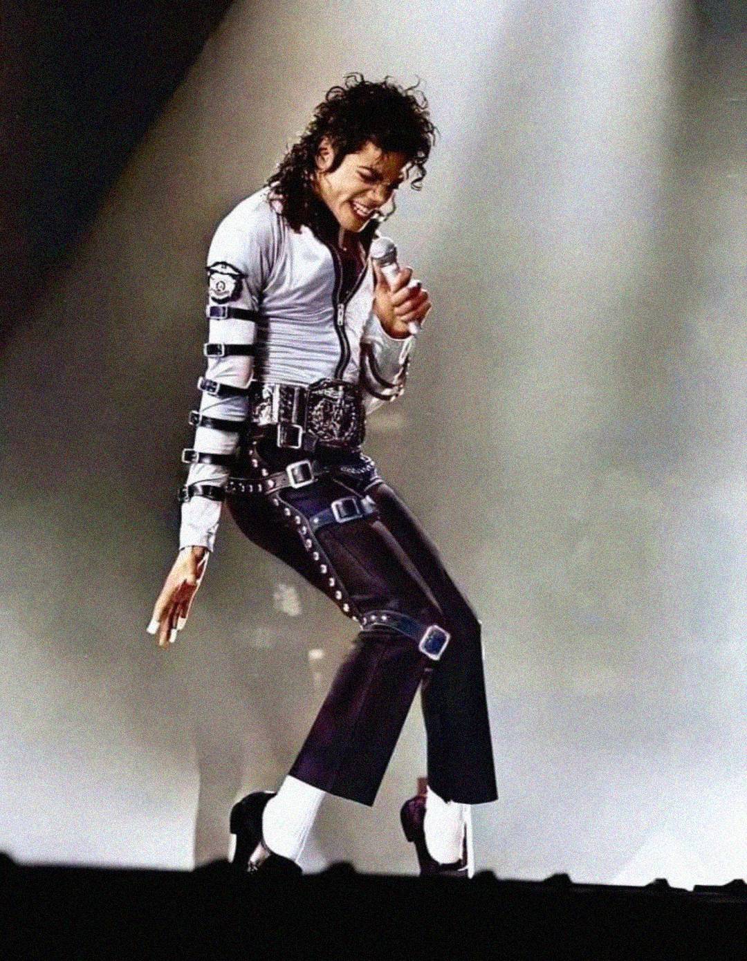 A performer dressed in a white shirt, black pants with buckles, and white socks, strikes a dance pose on stage with a spotlight behind them.