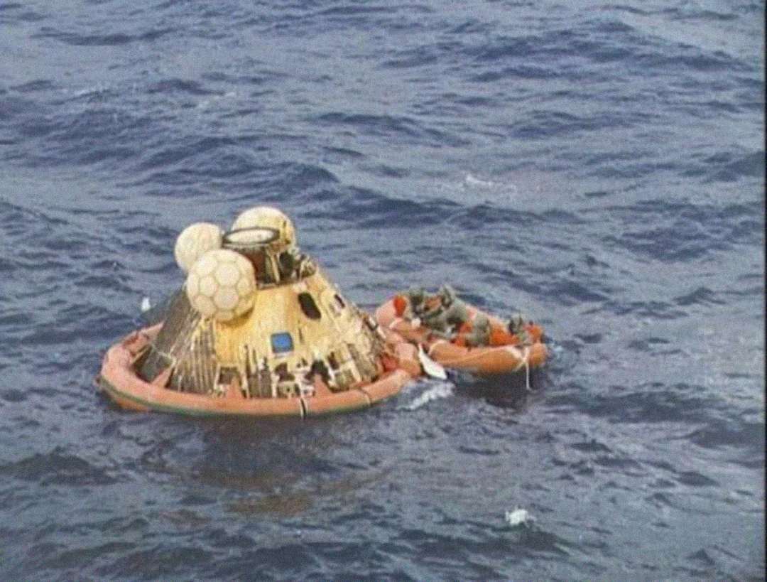 A space capsule floats in the ocean next to an inflatable orange life raft with several people aboard, likely astronauts and rescuers, following a splashdown landing.