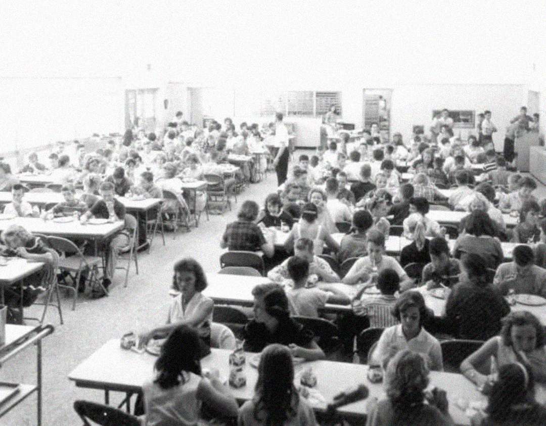 Black-and-white photo of a crowded school cafeteria with many children sitting at tables, eating and talking. The room is filled with natural light, and several adults are visible in the background near the serving area.