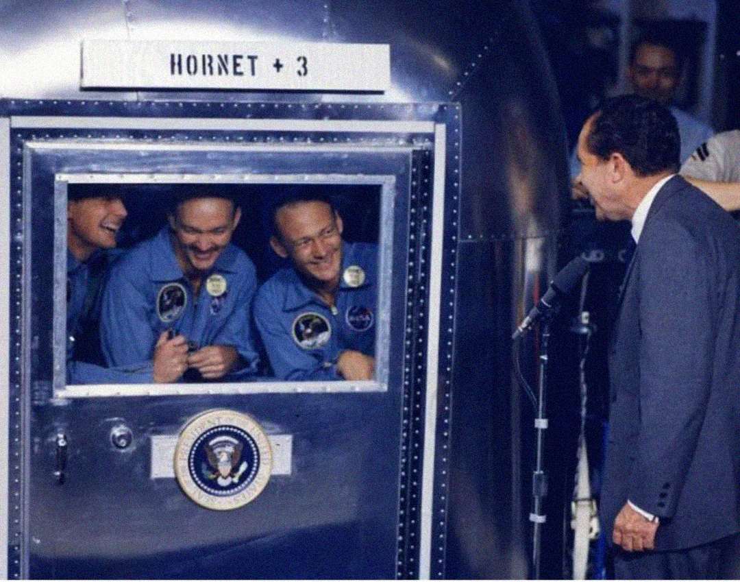 Three astronauts in blue suits smile and wave from inside a quarantine chamber labeled "Hornet + 3," while a man in a suit, speaking into a microphone, stands outside. The presidential seal is displayed on the chamber door.