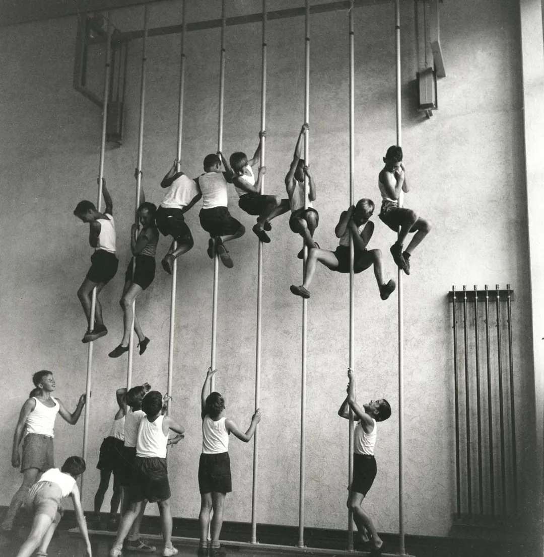 A group of children in gym clothes climb vertical poles in a gymnasium, while a few stand on the floor watching or waiting their turn. The image is black and white, with a plain wall in the background.
