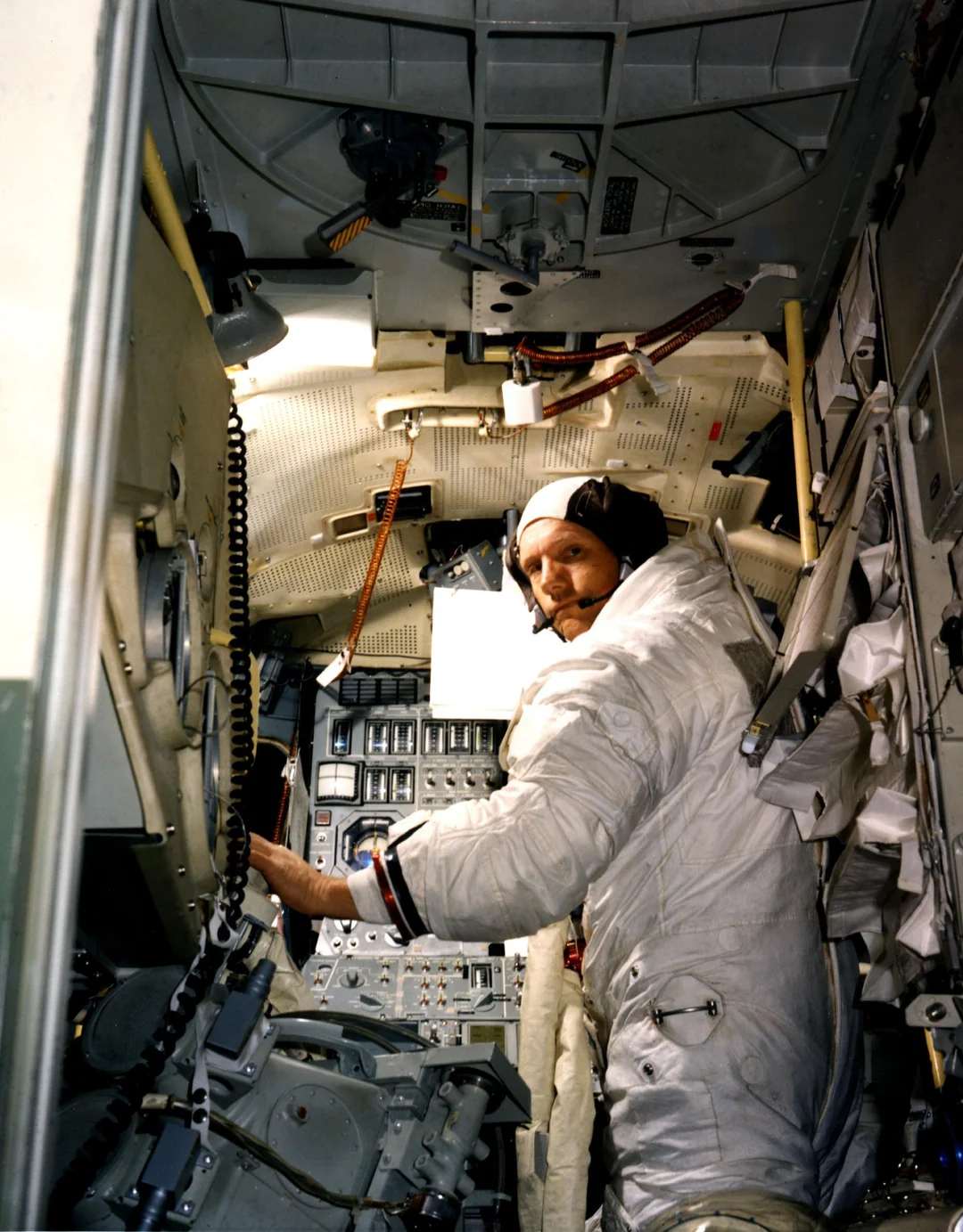 An astronaut in a white spacesuit and headset operates controls inside a cramped spacecraft cockpit filled with switches, buttons, wires, and technical equipment.