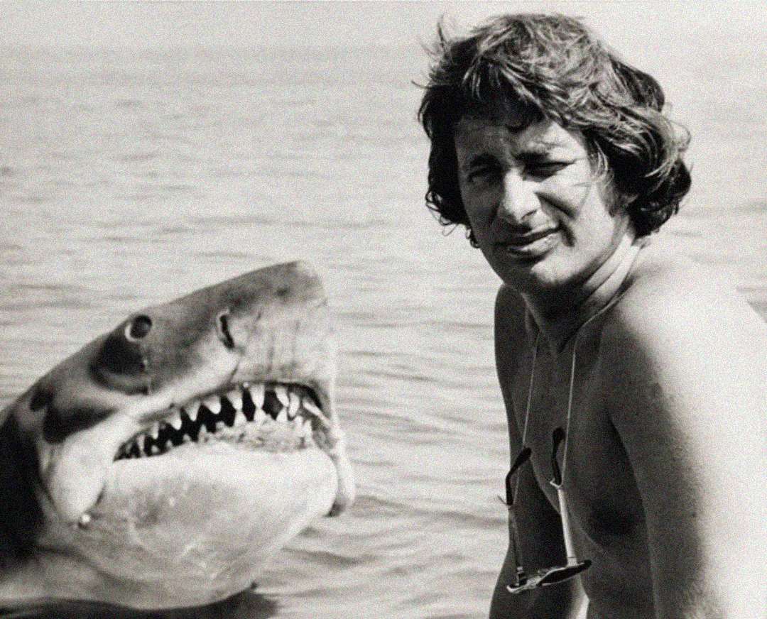 A black-and-white photo of a man with wavy hair, standing shirtless in the water, next to a large, lifelike mechanical shark with its mouth open, showing sharp teeth.