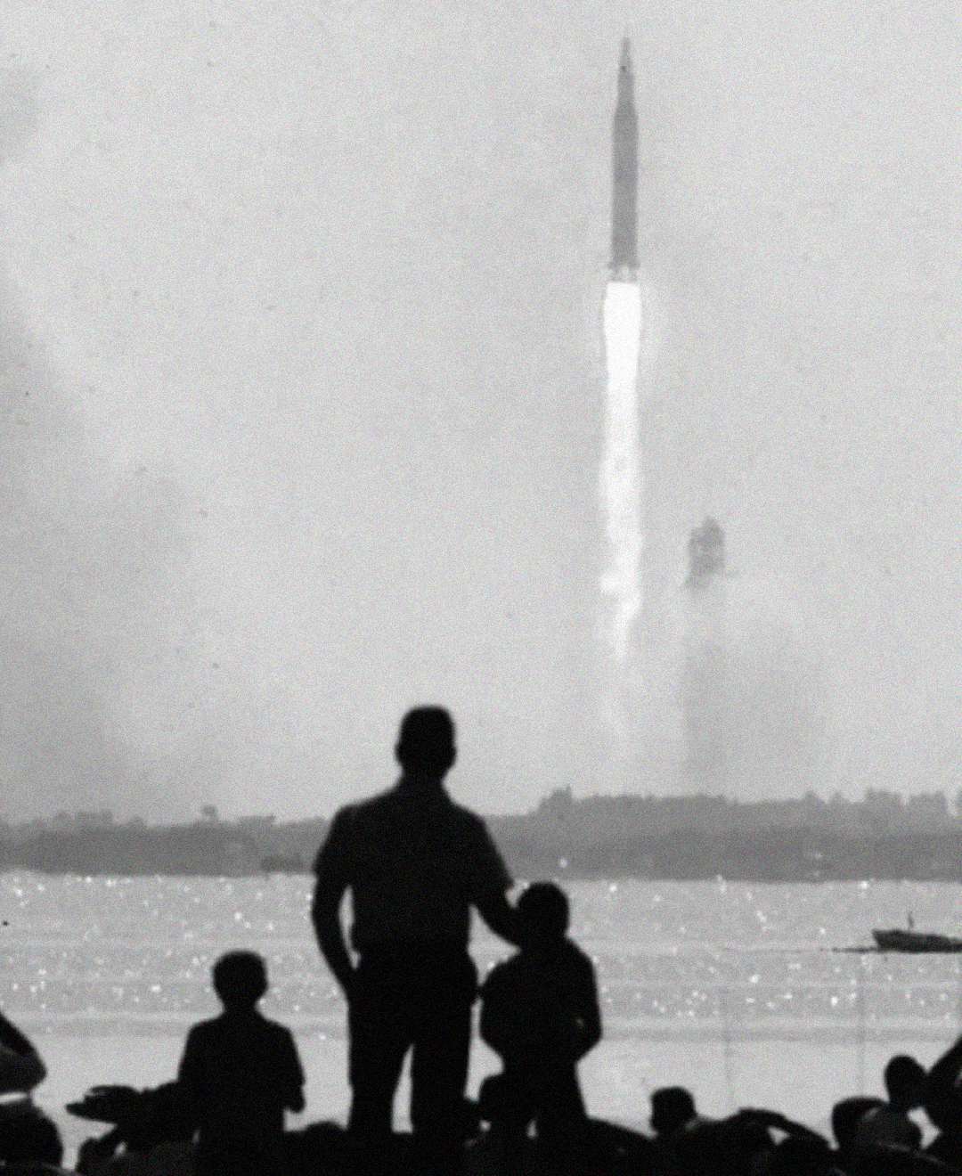 A black-and-white photo shows a rocket launching in the distance, with smoke trailing behind, while silhouetted people, including an adult and children, watch from across a body of water in the foreground.