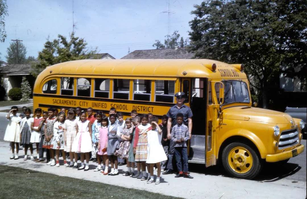 A group of schoolchildren and a uniformed driver stand in front of a yellow Sacramento City Unified School District bus on a sunny day in a residential neighborhood.