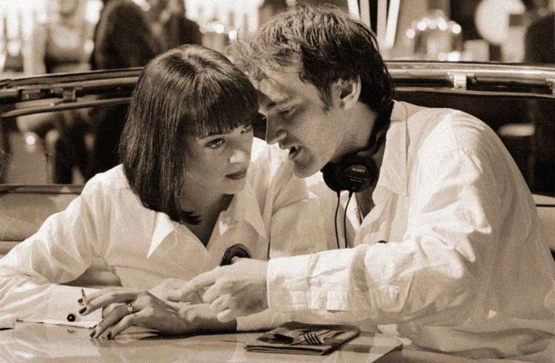 A woman with a bob haircut and a man wearing headphones around his neck sit closely together at a table, both dressed in white shirts, engaged in conversation in a retro setting.