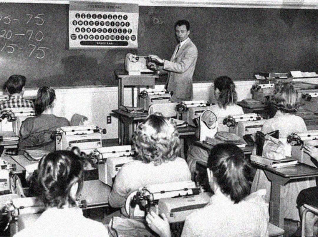 A black-and-white photo of a classroom shows a male teacher instructing students, who are seated at desks with typewriters. The teacher points to a chalkboard with numbers written on it.
