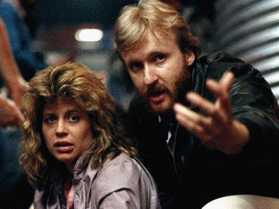 A woman with wavy hair looks tired while a bearded man beside her gestures with his hand, both appearing focused in a dimly lit, indoor setting.