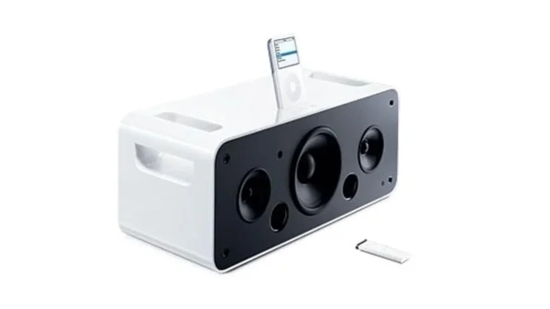 A white speaker dock with a black front panel, featuring multiple speaker drivers, an iPod inserted on top, and a small remote control placed nearby on a white background.