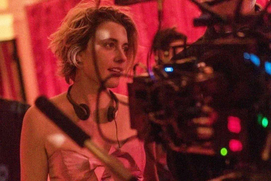 A person with short, tousled hair and headphones around their neck stands near film equipment, with pink lighting and a blurred background suggesting a film set.