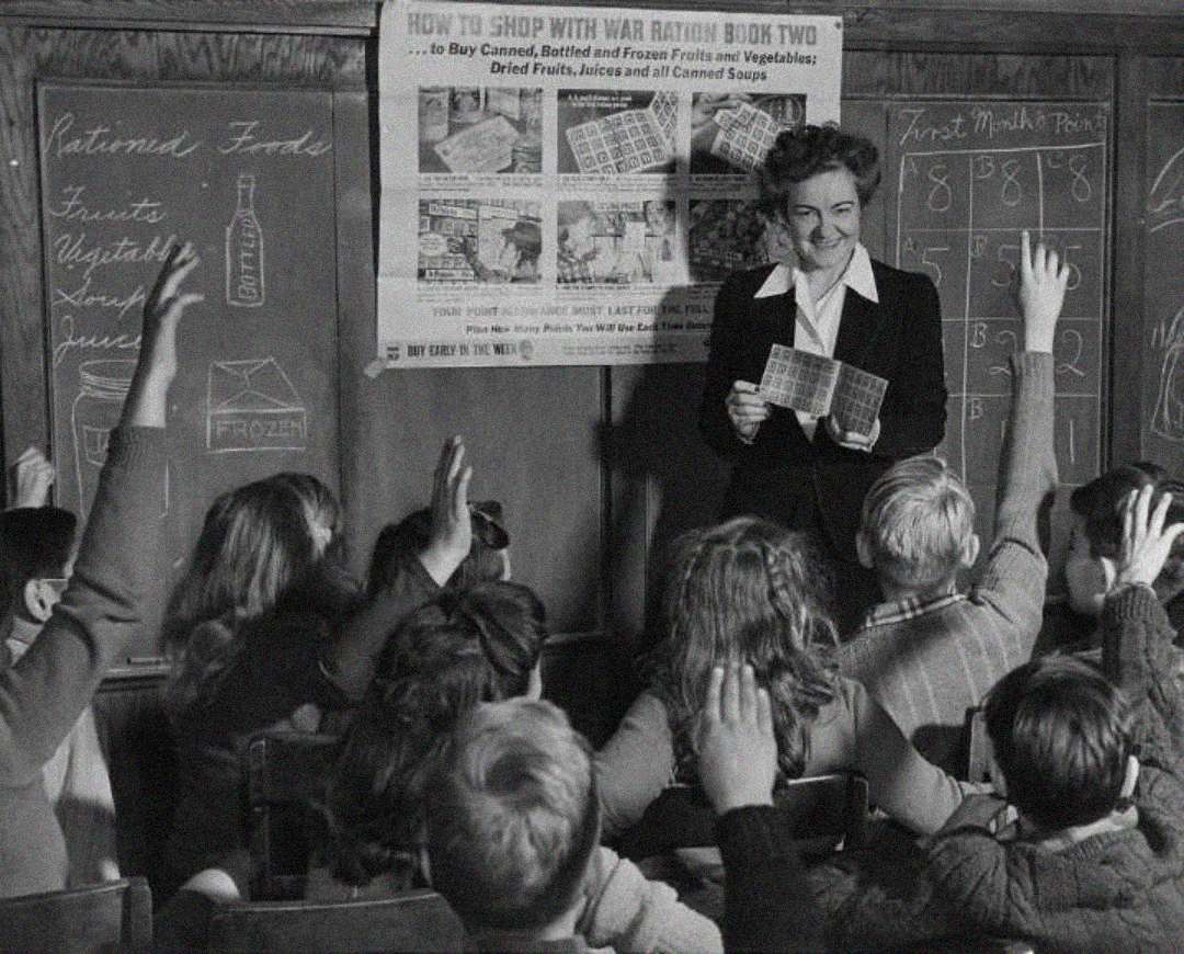 A woman stands in front of a classroom holding ration books, teaching children about wartime rationing. Several students sit at desks with hands raised. Chalkboards and a rationing poster are visible behind her.