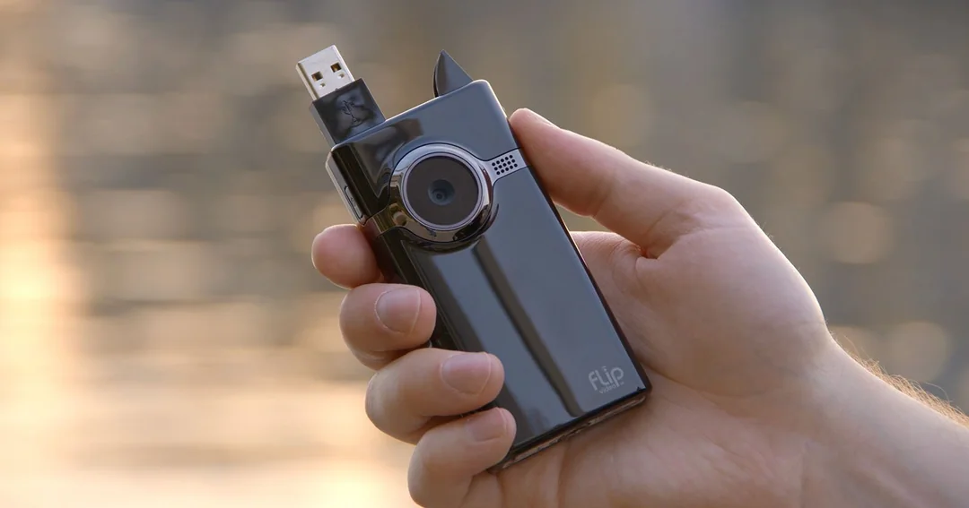 A hand holds a shiny black Flip video camera with its built-in USB connector extended. The camera lens and small microphone holes are visible on the front.