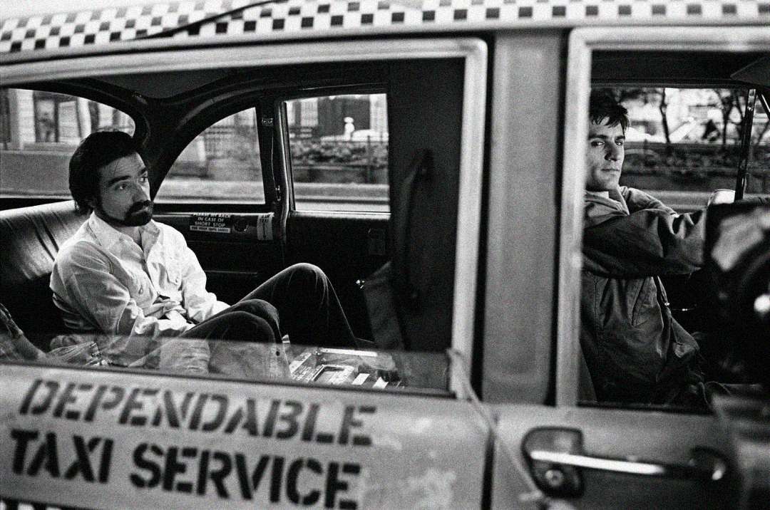 Black-and-white photo of two men inside a taxi; one sits in the back seat looking out thoughtfully, while the other, in the front driver’s seat, looks ahead. The taxi door shows "Dependable Taxi Service" text.