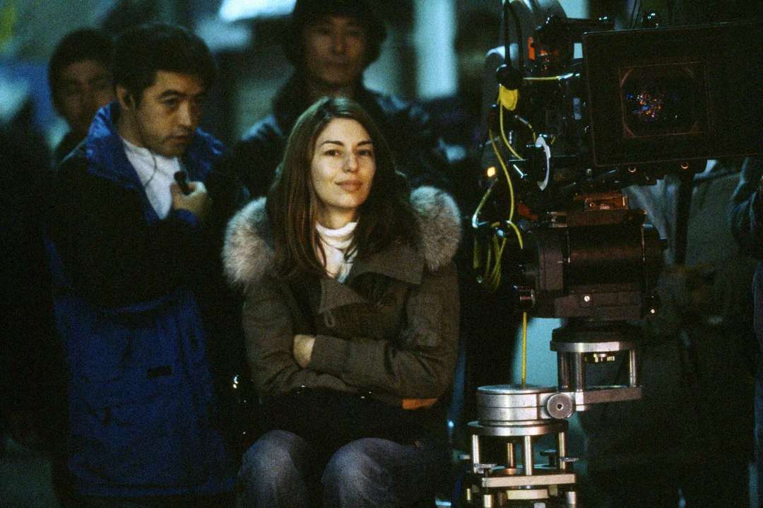 A woman in a winter coat with a fur-lined hood sits with her arms crossed on a film set, facing a large camera, while crew members stand around her in the background.