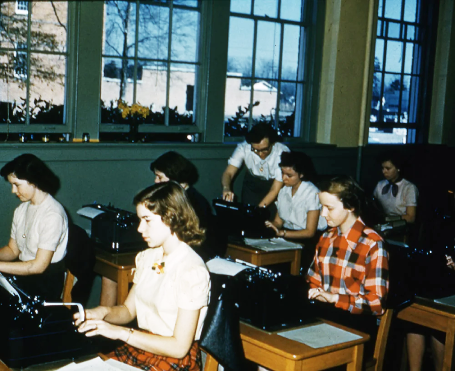 A classroom with students seated at desks using typewriters, while an instructor stands and assists. Large windows let in natural light, and trees are visible outside. The scene appears to be from a past decade.