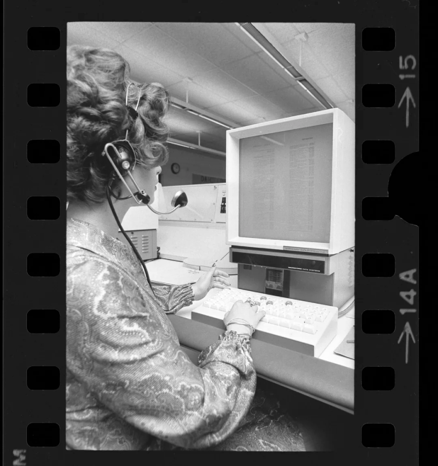 A woman wearing a headset types on a vintage computer with a large monitor in an office setting. The photo has a black film border with numbered frames visible.