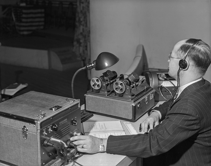 A man in a suit operates vintage audio recording equipment at a desk. He wears headphones and sits beside reels, a microphone, and a lamp, with a notepad and pen in front of him.