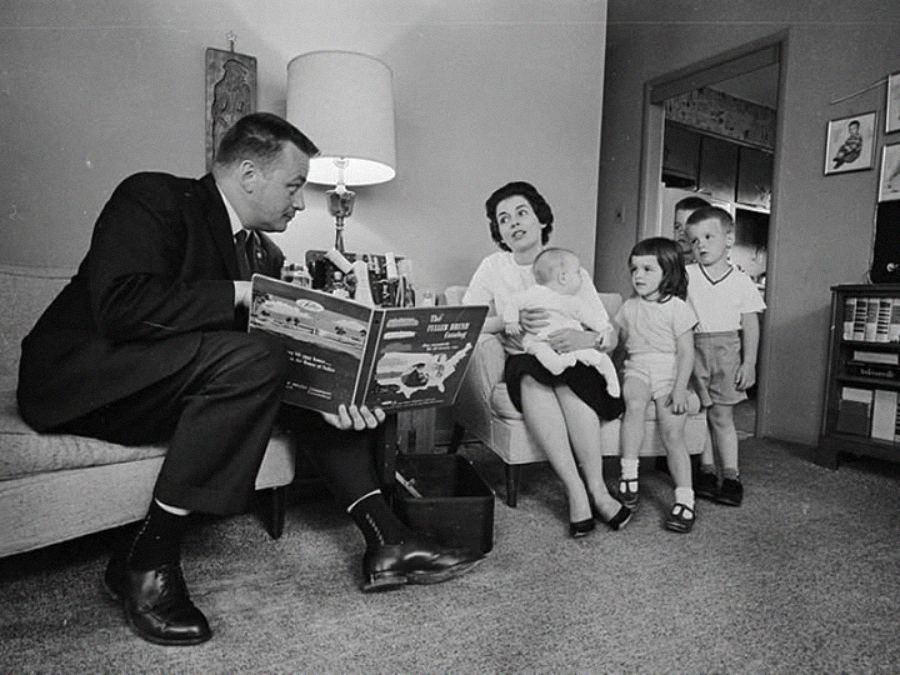 A man sits on a couch reading a book to a woman holding a baby on her lap, while two young children stand nearby listening in a cozy living room.