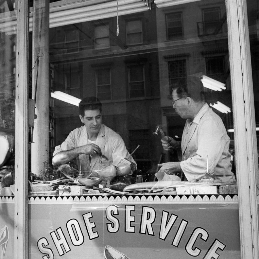 Two men work on shoes at a cluttered workbench inside a shoe repair shop, seen through a window with a sign reading “SHOE SERVICE.” Reflection of buildings and lights is visible on the glass.