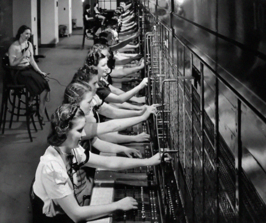 A row of women in vintage clothing operate a large telephone switchboard, plugging and unplugging cables, while another woman sits and speaks into a telephone in the background. The scene appears to be from the early to mid-20th century.