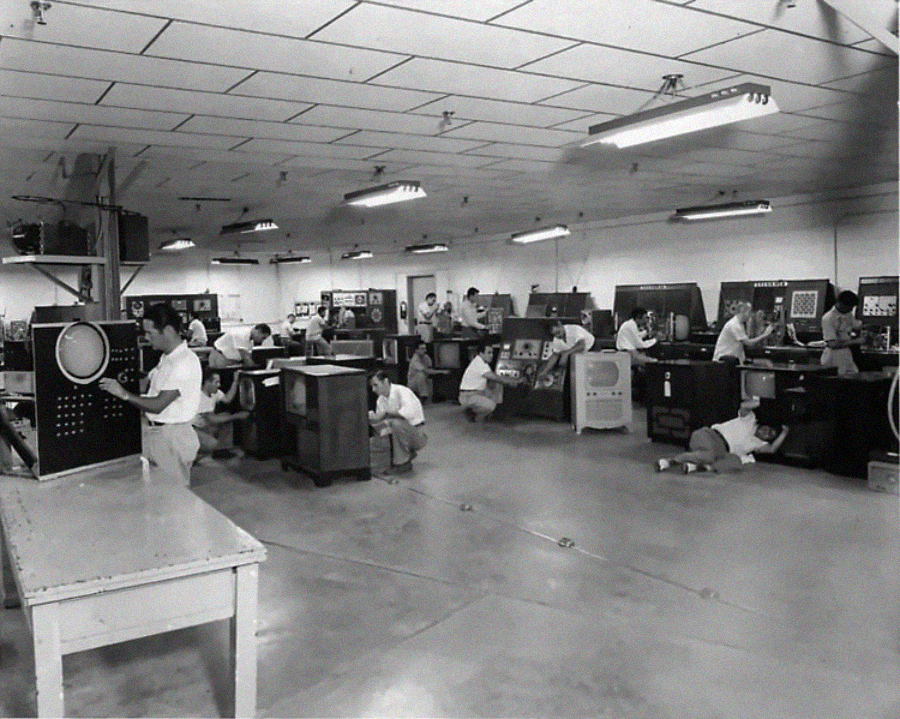 A group of people work on large, early computer machines in a spacious, fluorescent-lit room with a tiled ceiling. Some are seated or kneeling, interacting with control panels and screens.